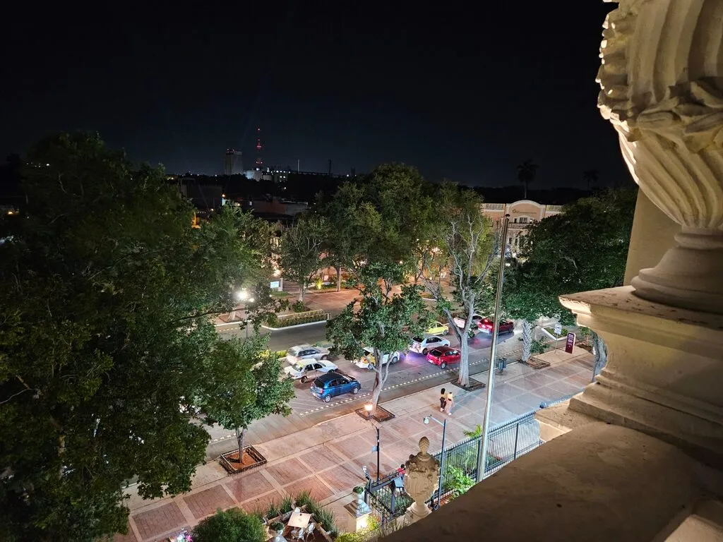 A tree-lined avenue at night seen from a carved stone balcony, with cars passing below.