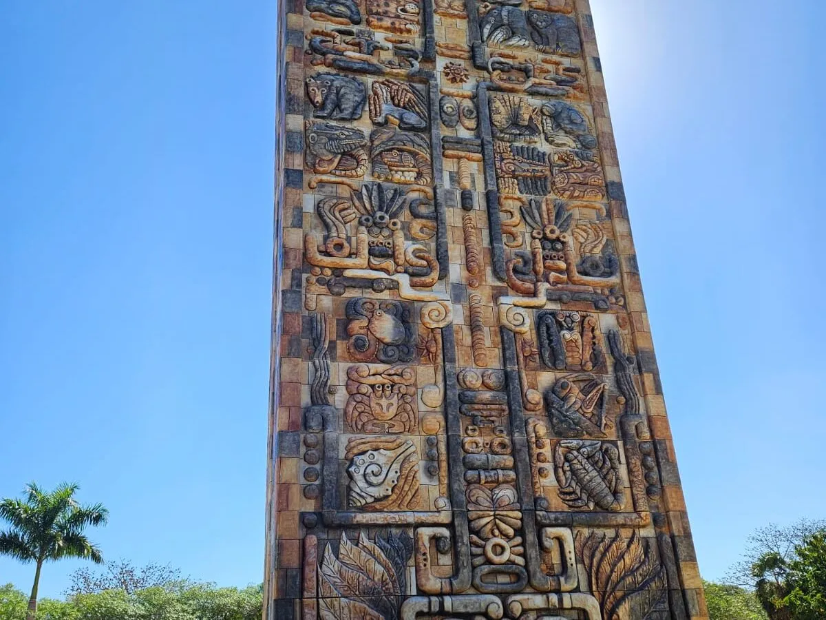 A tall monument carved with Maya-style reliefs against a bright blue sky, palms at its base.